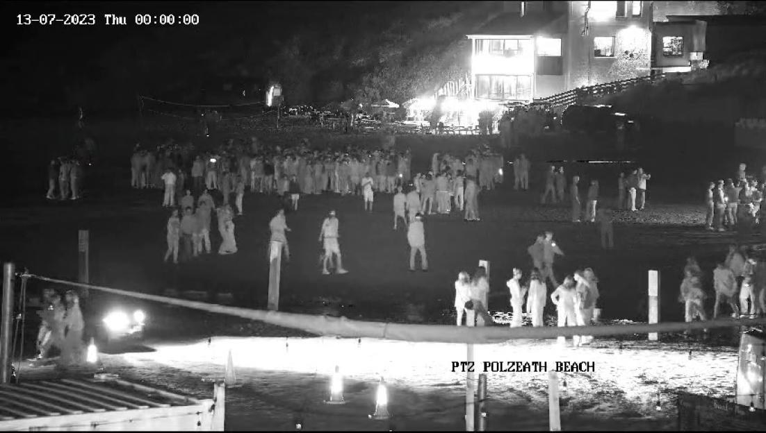 Black and white photo of a large crowd of people gathered on Polzeath beach at night, with buildings lit up in the background.