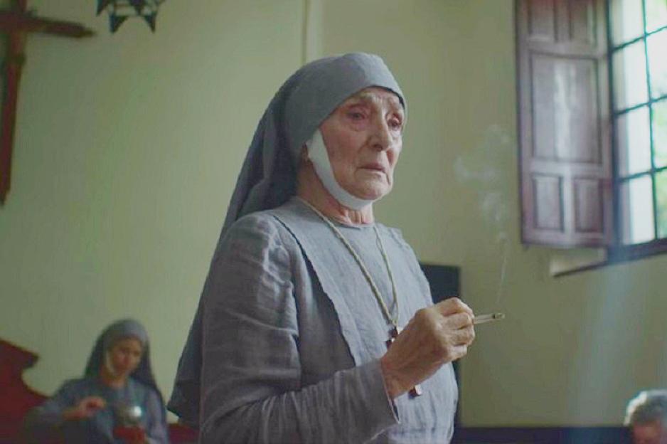 An elderly nun in a gray habit smokes a cigarette, with smoke curling from it, inside a room with a cross on the wall and a window.