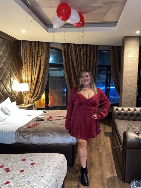 Becki Jones posing in a decorated hotel room with red and white balloons and rose petals on the bed.