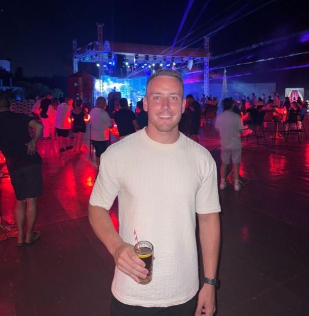 A man at an outdoor party at night holding a drink, with a crowd and stage with colorful lights behind him.