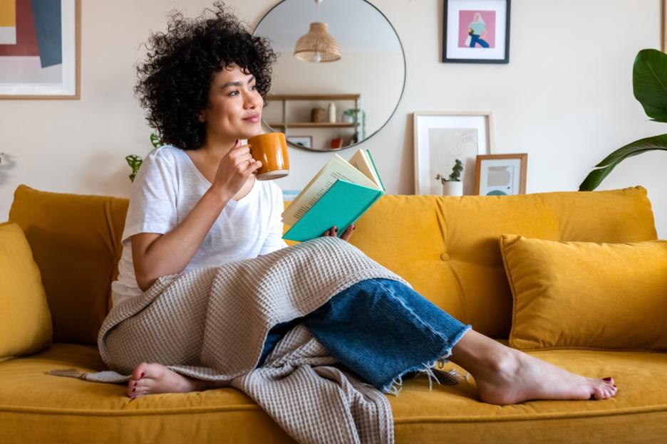 African American woman reading a book and drinking coffee on a yellow couch.