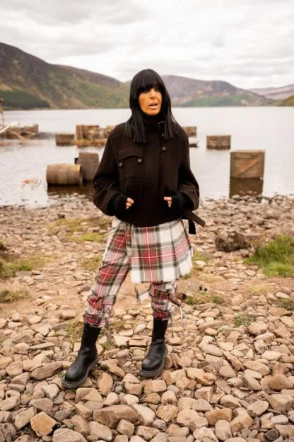 Claudia Winkleman standing by a loch in Scotland, wearing a dark jacket, plaid trousers, and a plaid kilt, with black boots.