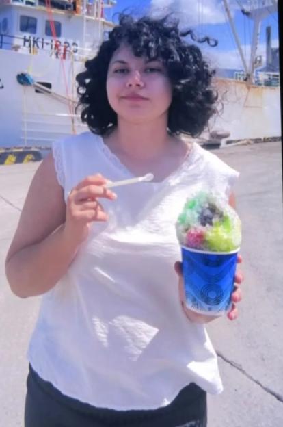 A woman stands with a cup of shaved ice in front of a white ship.