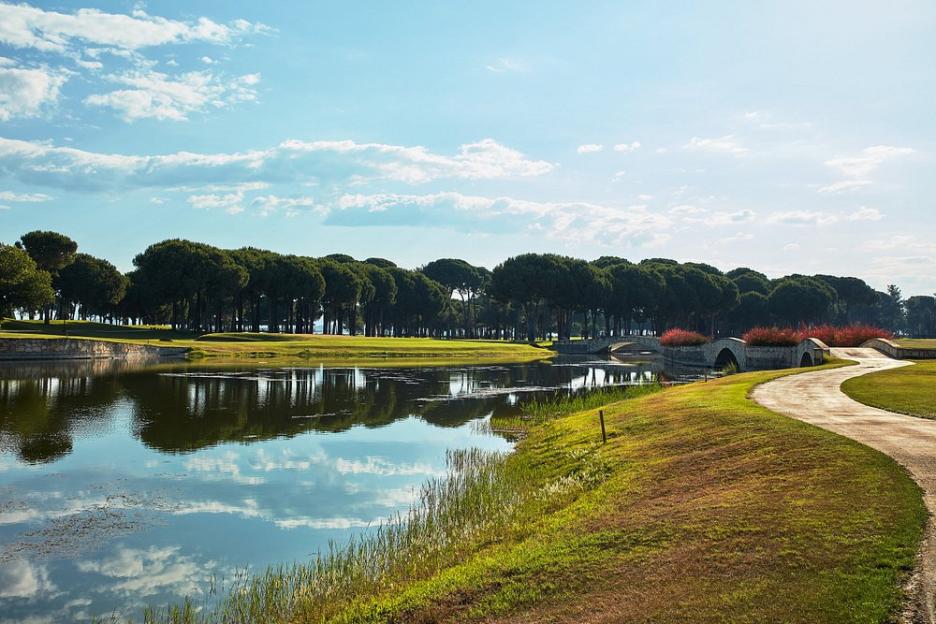View of Gloria Golf Resort's golf course with a pond, a bridge, and trees under a cloudy sky.
