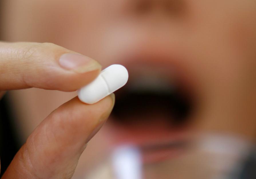 A person's fingers holding a white pill with a mouth open in the blurry background.