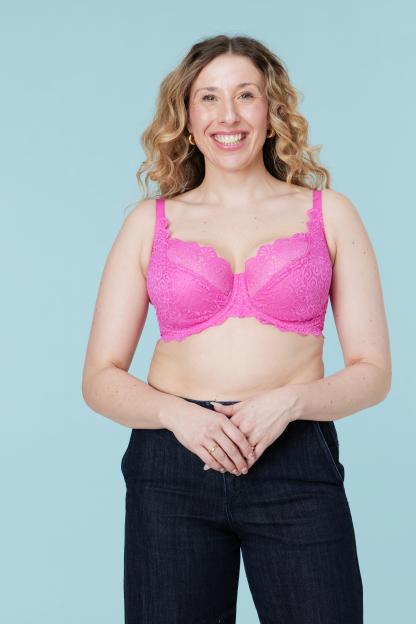 A smiling woman with curly hair and a pink lace bra, wearing dark jeans, stands against a light blue background.