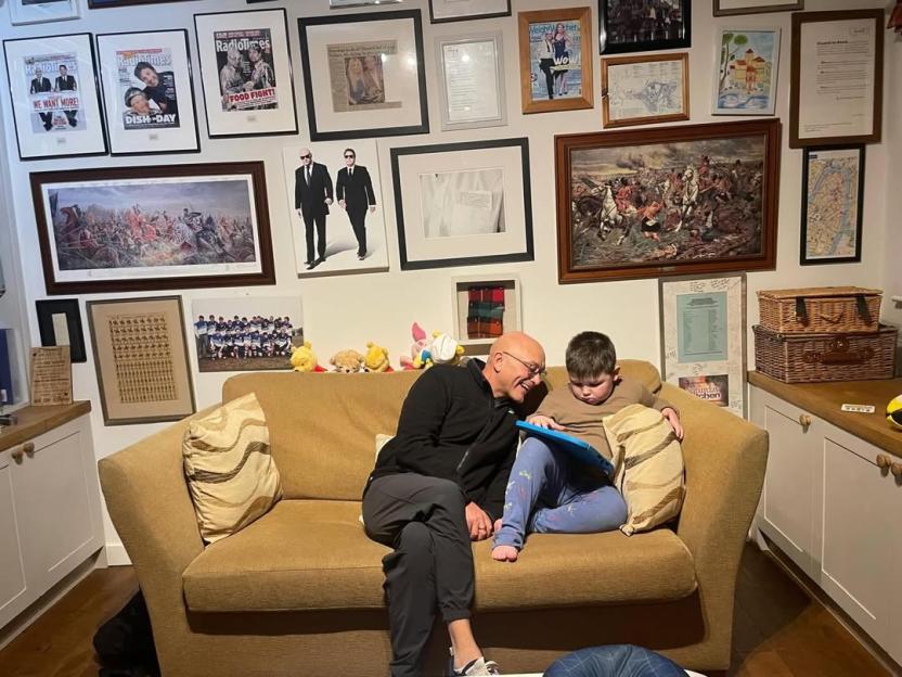 Gregg Wallace and his son Sid sitting on a sofa with many framed pictures on the wall behind them.
