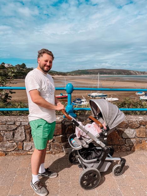 A man with a stroller standing next to a stone wall overlooking a bay with boats on the sand.