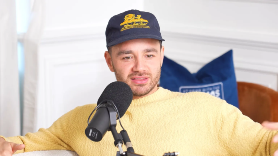 Man in a yellow sweater and baseball cap speaking into a microphone.