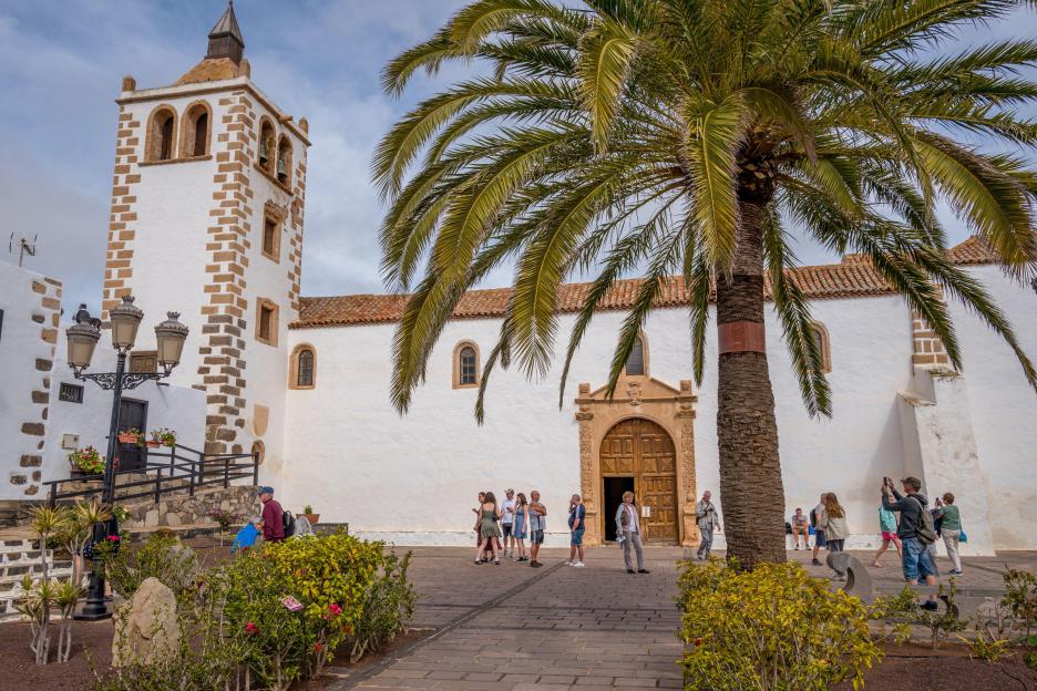 The Church of Santa María de la Concepción, a white building with a stone bell tower, with tourists and palm trees in the foreground.