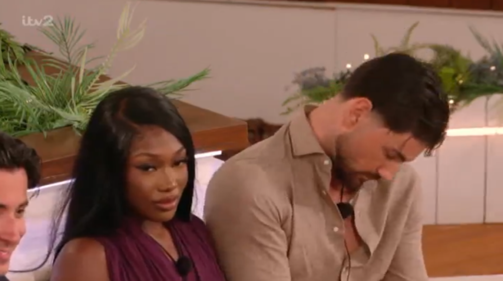 A woman in a purple top looks at the camera while a man next to her looks down.