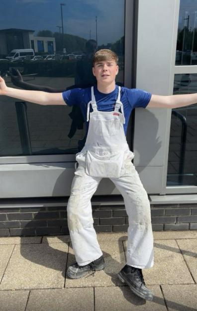 Kayden Moy, a 16-year-old in paint-splattered white overalls and a blue shirt, leans against a building.