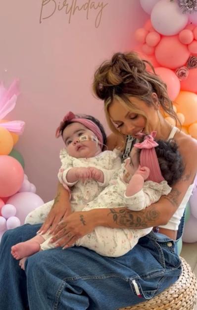 Jesy Nelson holding her twin babies who are wearing pink headbands and floral rompers.