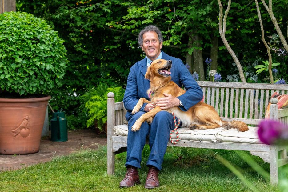 Monty Don sits on a wooden bench holding his golden retriever, Ned, in a garden.