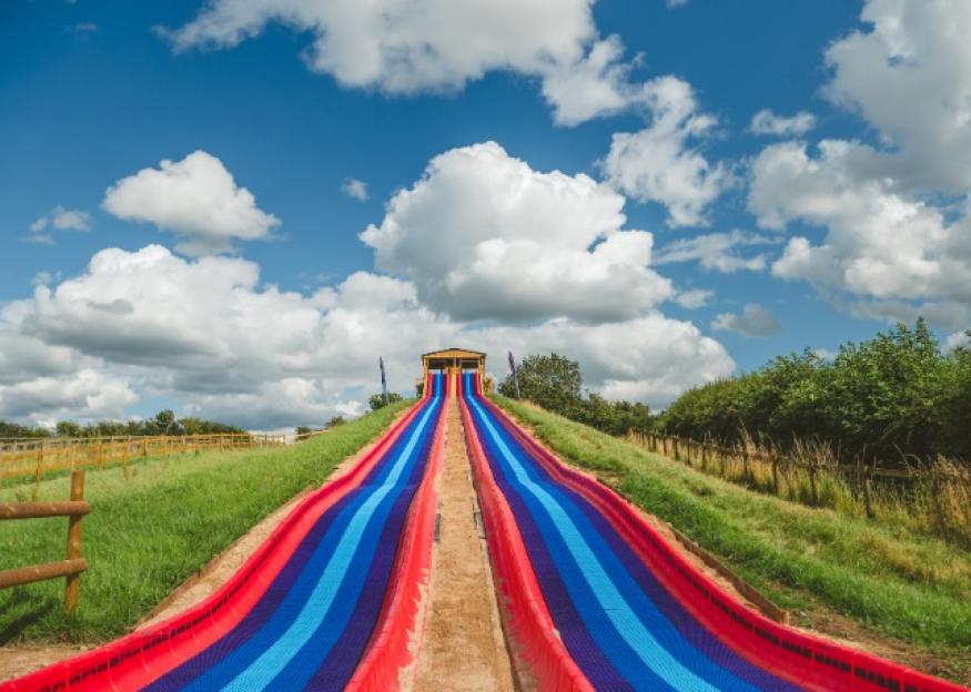 A long, colorful double slide with blue and red stripes stretching up a grassy hill under a blue sky with white clouds.