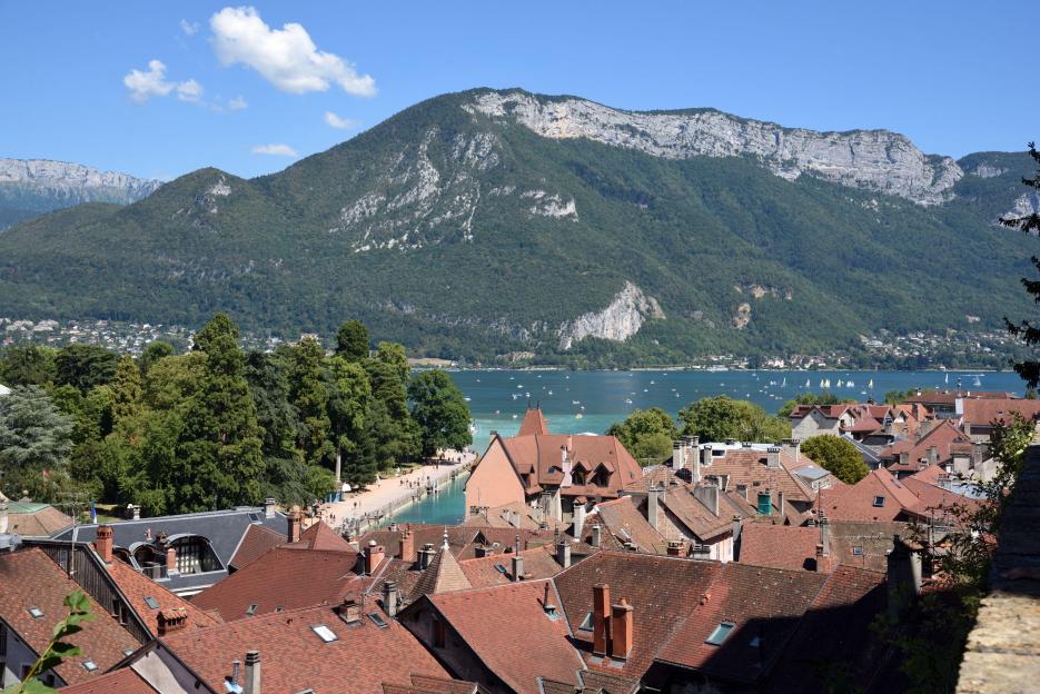 View over the red-tiled rooftops of Annecy's Old Town, with Lake Annecy and forested mountains in the background.