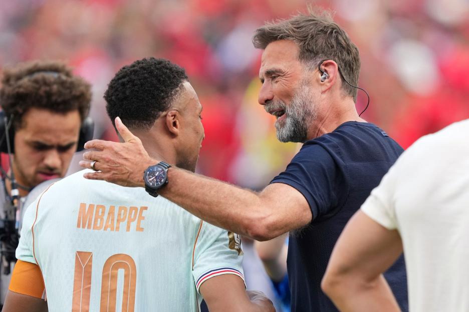 Kylian Mbappé in a France soccer jersey being embraced by a man in a navy shirt with a gray beard.