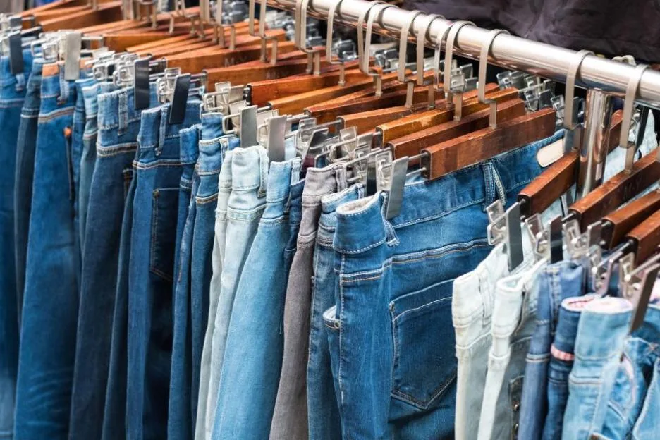 A row of various blue jeans hanging on wooden hangers at a market.