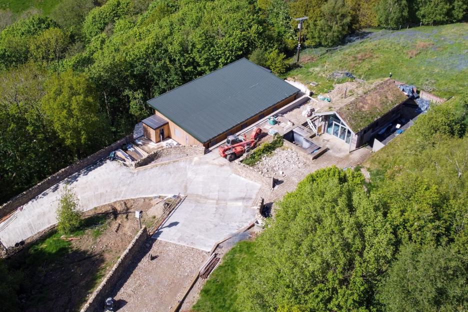 Aerial view of a farm in a forest clearing showing new construction, including a building with a green roof, a red tractor, and concrete driveways, representing groundworks for holiday homes.