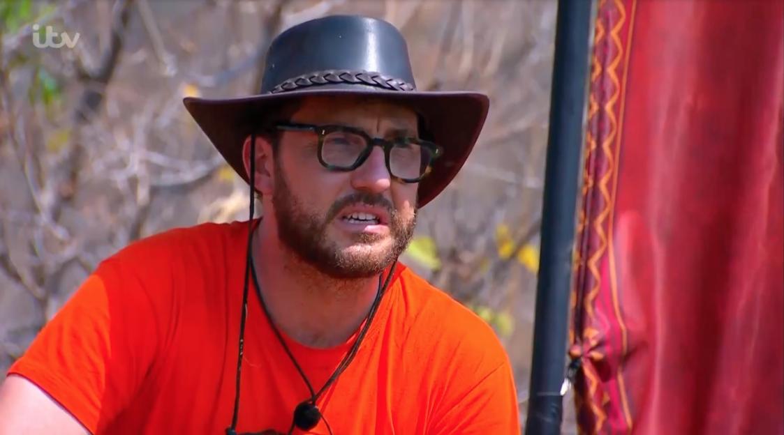 A man wearing a black hat, glasses, and an orange shirt looks to the right.