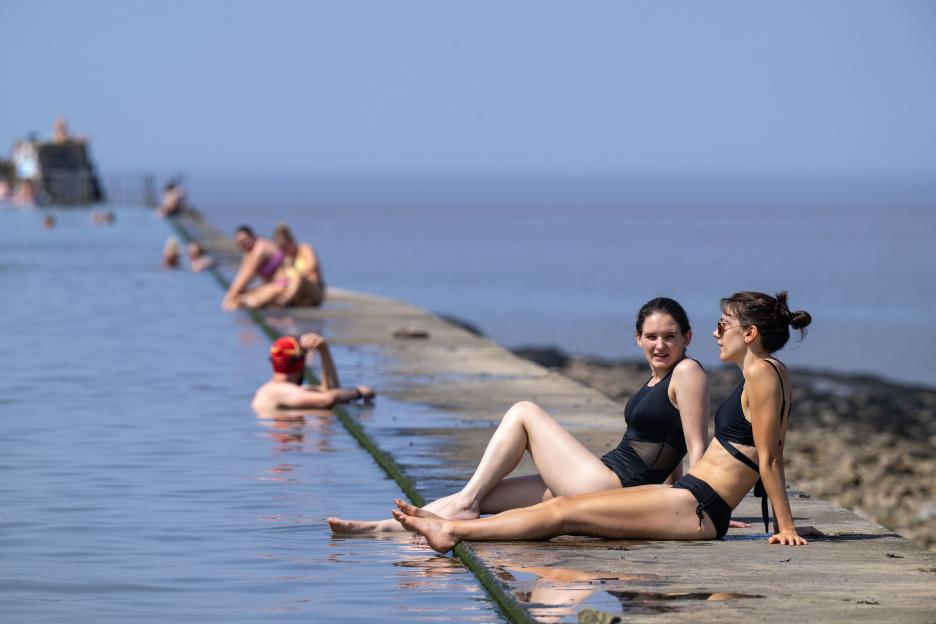 Two women sunbathe by the water, with other people swimming in the background.