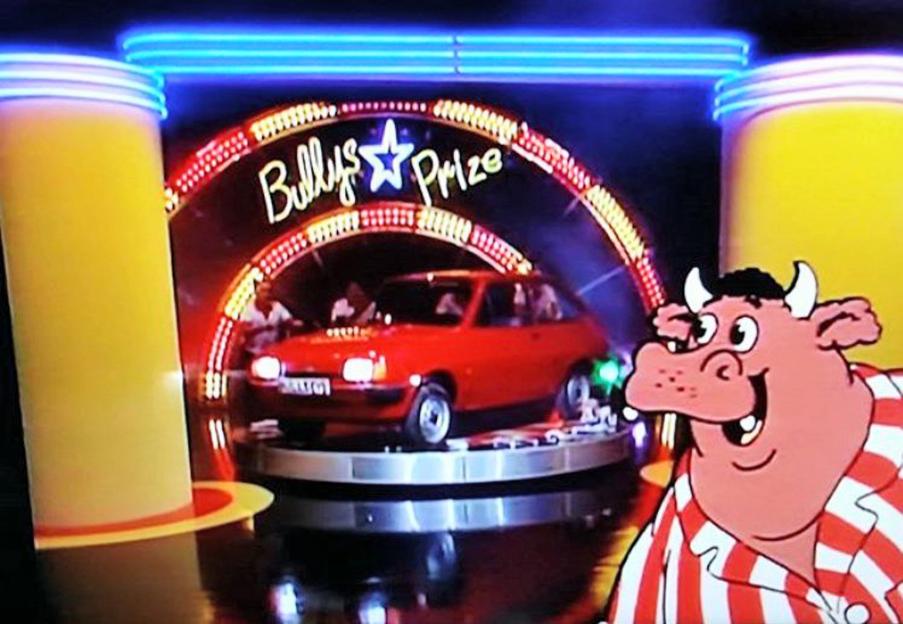 Bully, the anthropomorphic bull mascot, standing in front of a red Ford Fiesta on a rotating platform and a "Bully's Star Prize" sign from the game show Bullseye.
