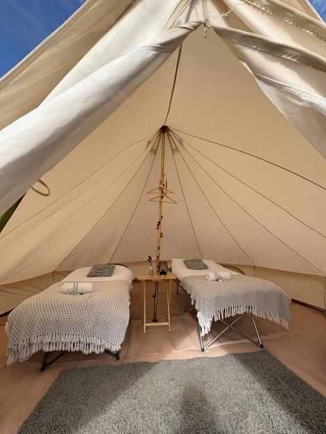 Inside a bell tent, two single beds with patterned blankets and pillows flank a small wooden table with a vase of flowers, a lantern, and a central pole decorated with lights and wooden hangers.
