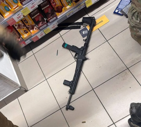 An assault rifle lies on a tiled floor in a supermarket aisle, with chocolate bars on shelves in the background.