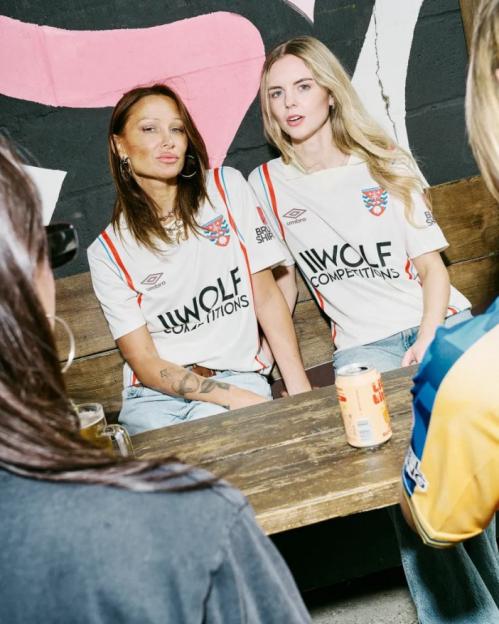 Two women in white football jerseys with the "WOLF COMPETITIONS" logo sitting at a wooden table.