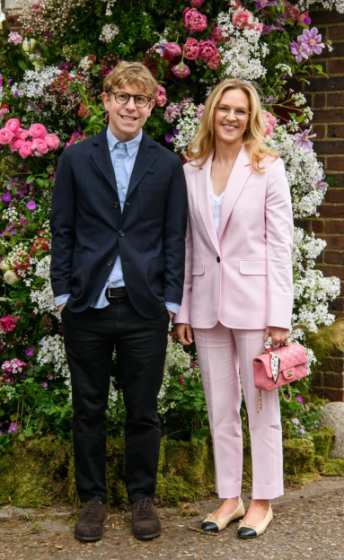 A man in a navy suit and a woman in a pink suit pose in front of a flower arrangement.