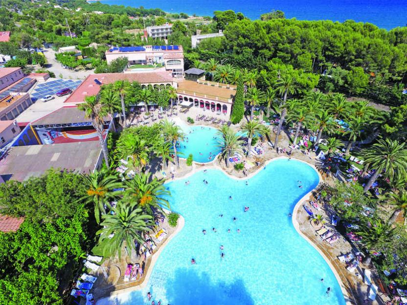 Aerial view of a resort with two large swimming pools, surrounded by palm trees and buildings with a coastal view in the background.