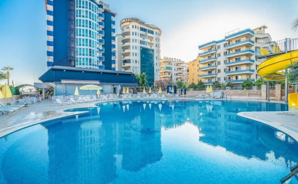 A large swimming pool surrounded by a patio with lounge chairs and yellow umbrellas, with tall blue and white buildings in the background.