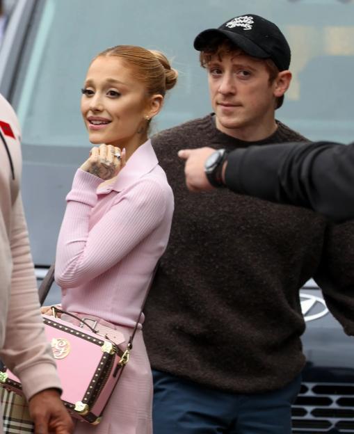 Ariana Grande, smiling in a pink shirt and skirt, and Ethan Slater in a dark sweater and cap, are shown arriving in Sydney.