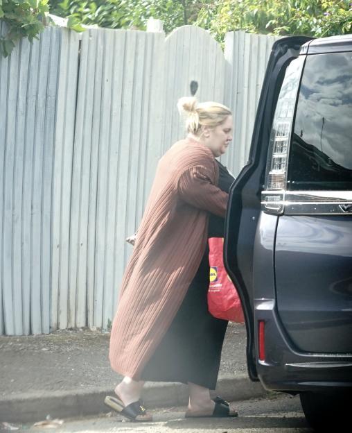 Woman in a brown cardigan and black slides getting out of a dark car, holding a red shopping bag.