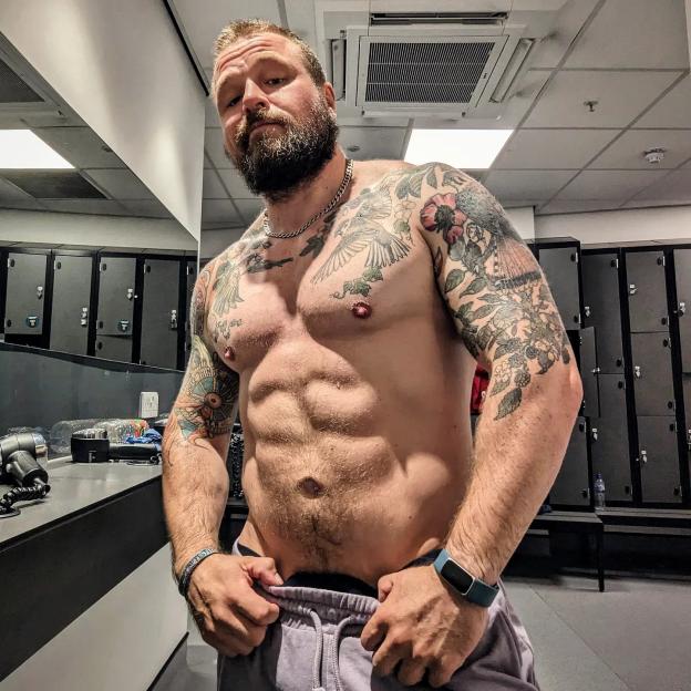 David Paisley shirtless, showing off his tattoos, muscles, and beard in a locker room.