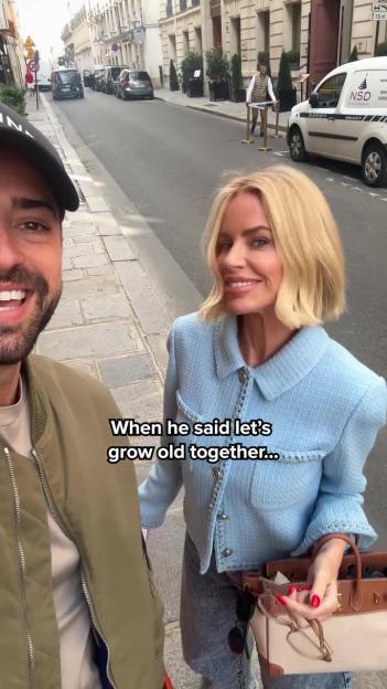 A smiling woman, Caroline Stanbury, next to a man taking a selfie on a city street.