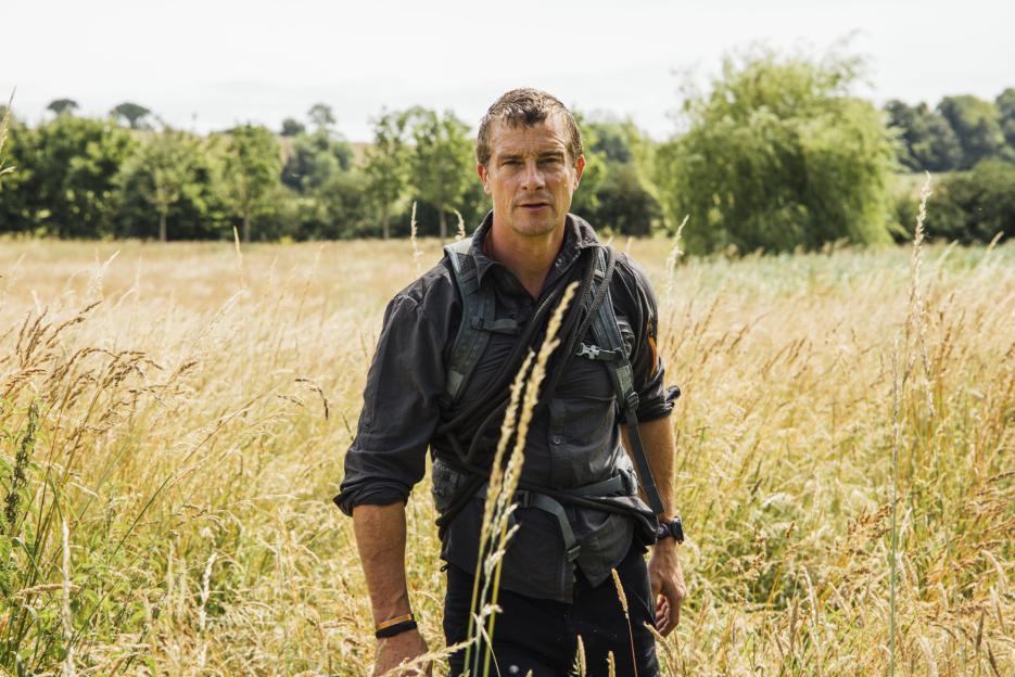 Bear Grylls standing in a tall, golden field with a backpack and rope, looking directly at the camera.