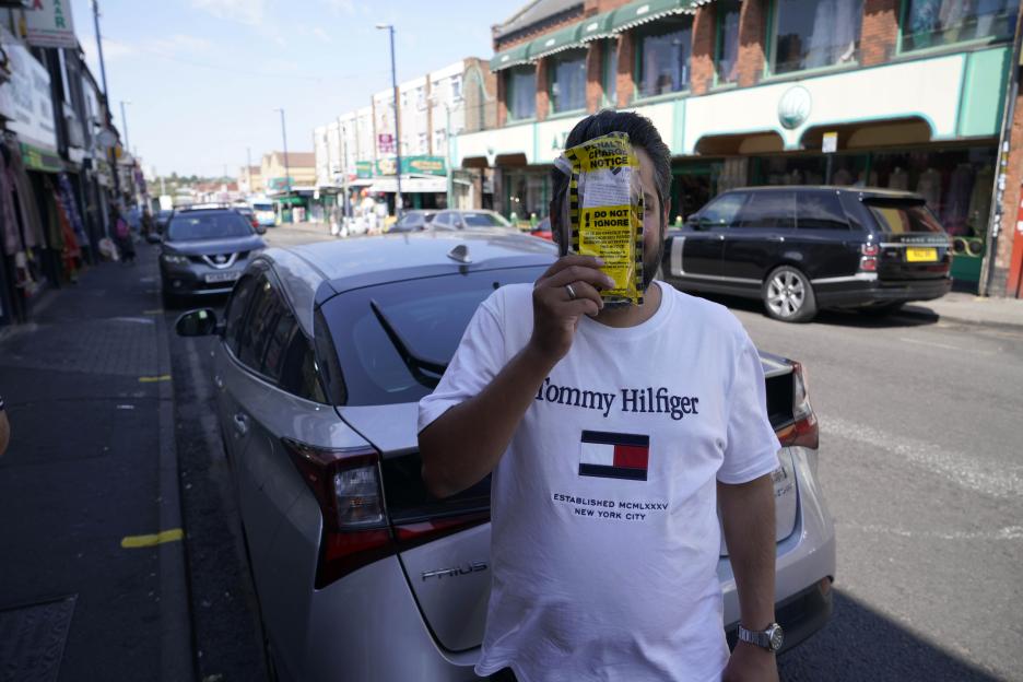A man holding a yellow parking ticket over his face next to a car, on a street known for parking chaos and fines.