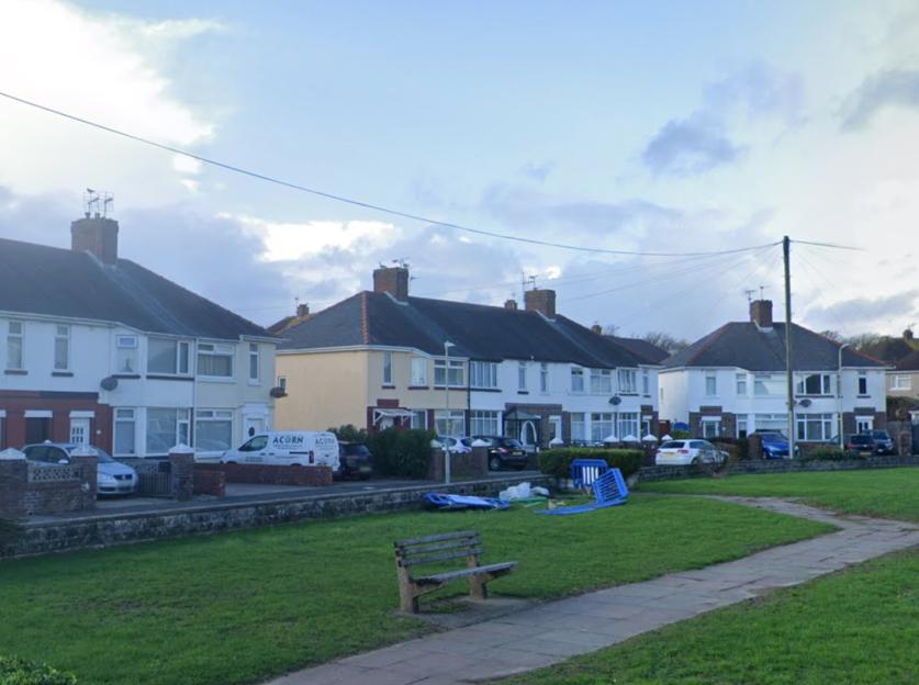 Street view of Poplar Crescent, Porthcawl, with multiple houses lining the street, a grassy area with a bench in the foreground, and cars parked along the street.