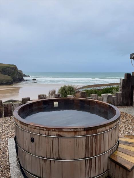 A wooden hot tub on a gravel patio overlooking a sandy beach and cliffs with an overcast sky.