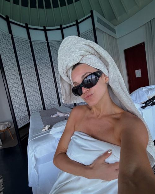 A woman in sunglasses with a towel on her head and wrapped around her body takes a selfie in a room.