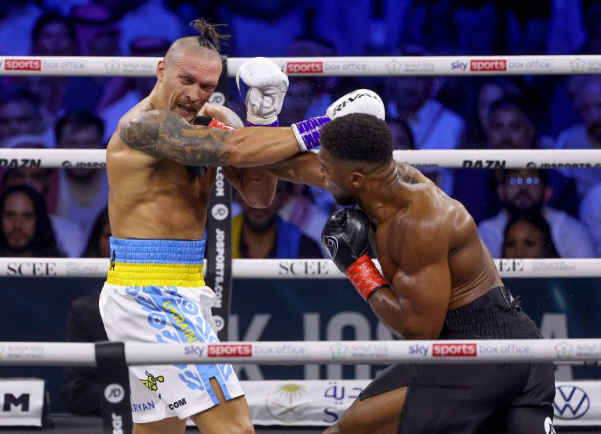 Boxer Anthony Joshua stands in the ring with sweat covering his torso, wearing white shorts and light brown boxing gloves, with "USYK" visible in the background.