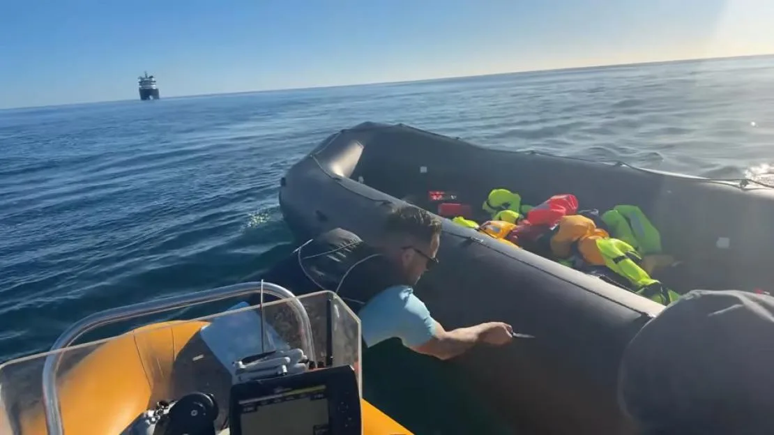 A man slashes an inflatable boat full of life jackets with a knife, with a large ship in the background.