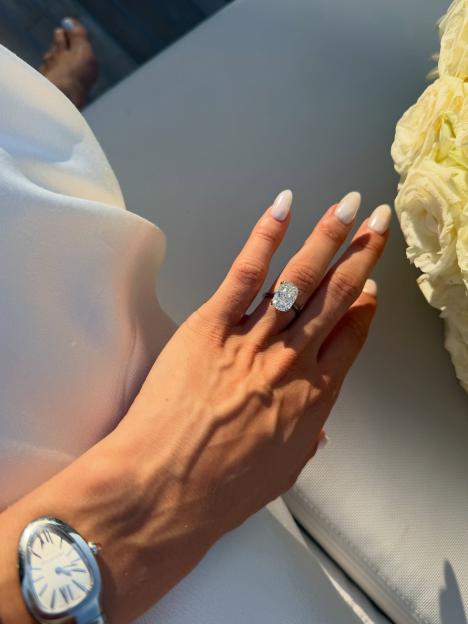 Nathalia Campos's hand with a large cushion-cut diamond engagement ring on her ring finger, a silver watch on her wrist, and a white flower in the upper right.