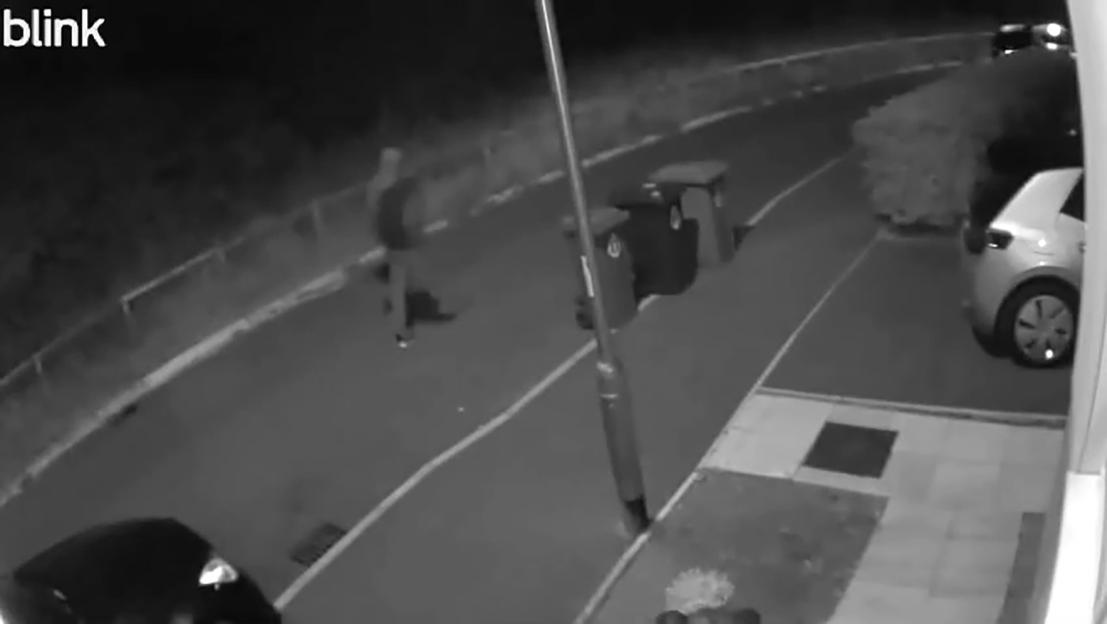 CCTV footage shows a person running on a sidewalk next to a road, with two parked cars, bushes, and two trash cans also visible.