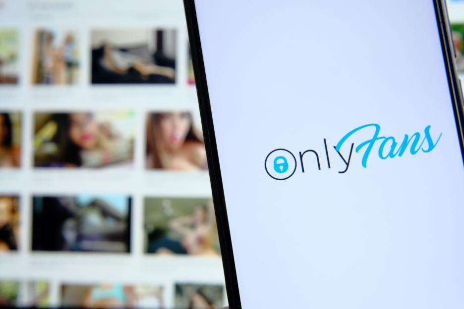 Onlyfans platform logo seen on smartphone and blurred pictures of unrecognisable girls on the background laptop. Site selling explicit pictures. Conce
