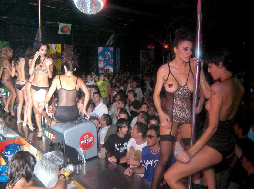 Several women in revealing outfits dancing on a bar and around a pole, with a crowd of men watching.