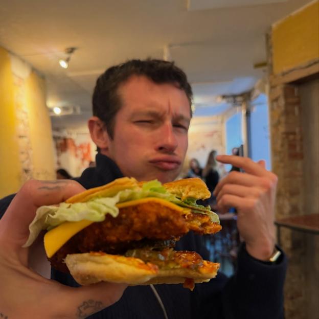Callum Turner holding a large chicken burger with cheese and lettuce.