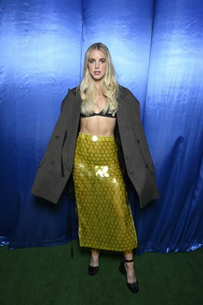 A person in a black bra, sequined yellow skirt, and oversized blazer poses for a photocall.