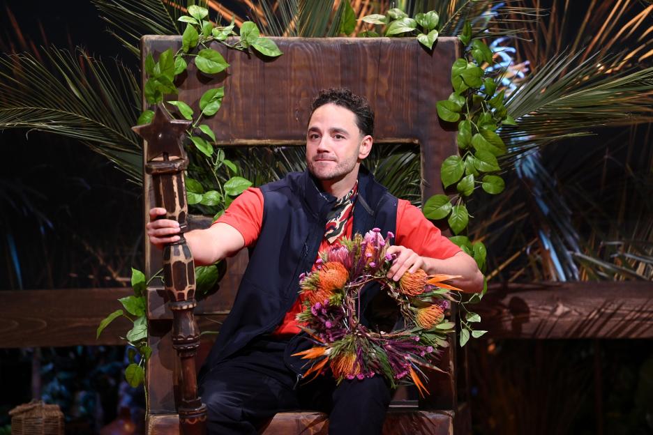 Adam Thomas sitting on a throne, holding a wooden scepter and a floral wreath.